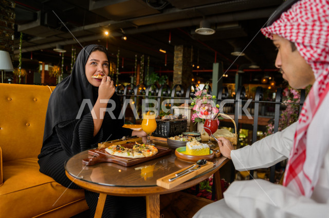 زوجان عربيان خليجيان سعوديان يجلسان على مائدة الطعام و يتناولان الطعام في المطعم ، قضاء اجواء رائعة  في المطعم ، تشكيلة متنوعة من أصناف الطعام و الماكولات الشهية على الطاولة ، جلسات فريدة و مميزة ، مطاعم و كافيهات في السعودية