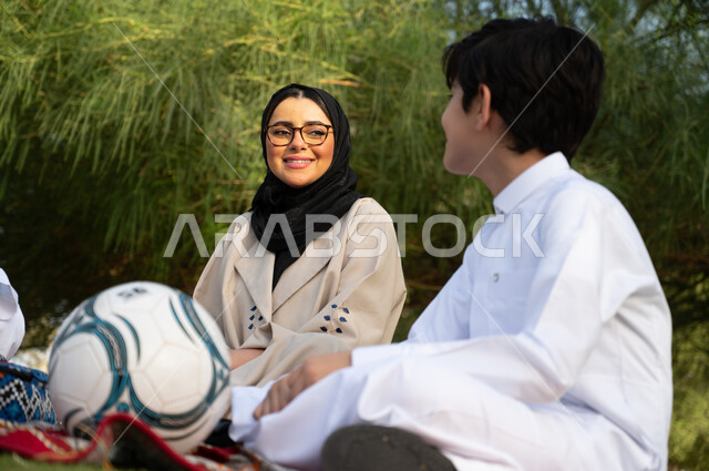 طفل عربي خليجي سعودي يجلس بجوار والدته ويقومان بتبادل النظرات, قضاء أوقات عائلية بالجلوس في الطبيعة الخضراء، الأجواء العائلية السعيدة, الحدائق العامة في المملكة العربية السعودية، نزهة في في الهواء الطلق، عطلة صيفية ترفيهية.