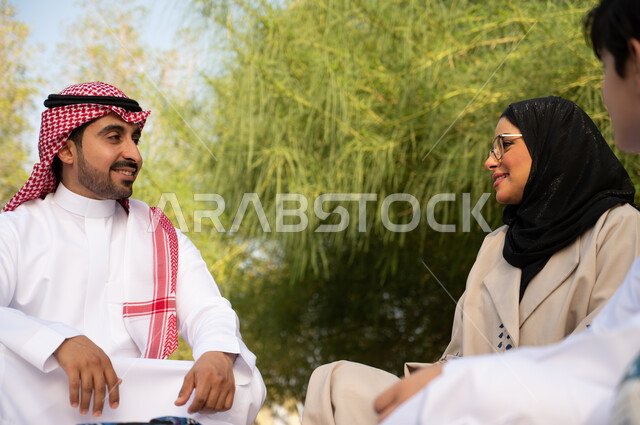 زوجان عربيان خليجيان سعوديان ينظران لبعضهما برفقة ابنهما في الطبيعة الخضراء في إحدى الحدائق العامة في المملكة العربية السعودية, قضاء أوقات ممتعة بالجلوس في الهواء الطلق، الأجواء العائلية السعيدة, عطلة صيفية ترفيهية. 