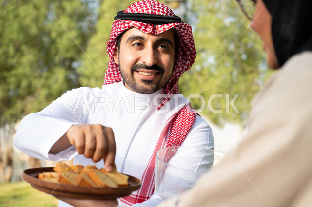 Eating pieces of cake, a Saudi Arabian Gulf couple in the green nature in one of the public parks in the Kingdom of Saudi Arabia, spending enjoyable times sitting in the open air, a happy family atmosphere, an entertaining summer vacation.