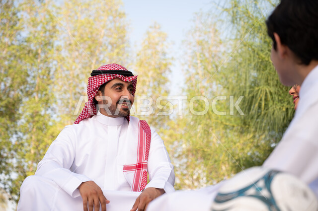 ولد عربي خليجي سعودي يجلس بجوار والده في إحدى الحدائق العامة في المملكة العربية السعودية، قضاء أوقات عائلية بالجلوس في الطبيعة الخضراء، الأجواء العائلية السعيدة, نزهة في في الهواء الطلق، عطلة صيفية ترفيهية.