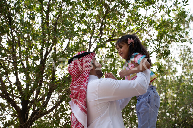 A Saudi Arabian Gulf father wearing a traditional shemagh and thobe carrying his little daughter in the open air, family parks and gardens, entertaining activities for children, spending enjoyable times in the green nature in one of the parks of the Kingd