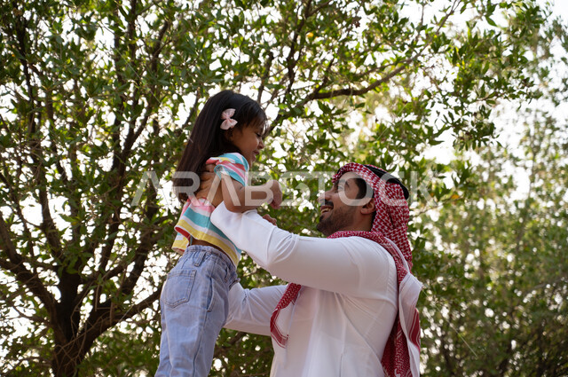 أب عربي خليجي سعودي يرتدي الشماغ و الثوب التقليدي يحمل ابنته الصغيرة في الهواء الطلق, منتزهات و حدائق عائليه, فعاليات ترفيهية مسلية للأطفال, قضاء أوقات ممتعة في الطبيعة الخضراء في إحدى حدائق المملكة العربية السعودية.