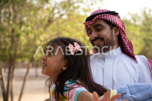 ايماءات وجه تدل على السعادة و السرور, أب عربي خليجي سعودي يرتدي الشماغ و الثوب التقليدي يحمل ابنته الصغيرة في الهواء الطلق, منتزهات و حدائق عائليه, فعاليات ترفيهية مسلية للأطفال, قضاء أوقات ممتعة في الطبيعة الخضراء في إحدى حدائق المملكة العربية السعودية.