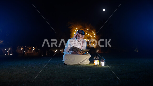 Lighting and decoration for the month of goodness, a Saudi Arabian Gulf man dressed in the traditional Saudi dress, holding the Holy Qur’an in his hand and sitting in the public garden of the house, reciting the Noble Qur’an, a spiritual atmosphere, Islam