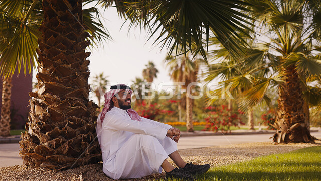 قضاء وقت ممتع في الطبيعة الخضراء وقت النهار, رجل عربي خليجي سعودي يرتدي الثوب والشماغ السعودي و يجلس تحت شجرةالنخيل , نزهه في الهواء الطلق, أماكن ترفيهية سياحية, التشجير وزراعه النخيل، ايماءات تدل على السعادة، خلفيه الطبيعه