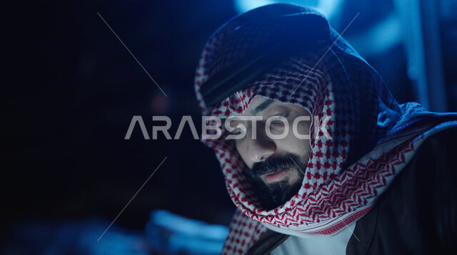 مخيمات سياحية في الصحراء، التواصل الاجتماعي عبر تطبيقات الجوال، صوة مقربة لرجل عربي خليجي سعودي يرتدي ملابس الشتاء الدافئة ويحمل بيده الهاتف المحمول مع اضاءة خافته ، التسوق الإلكتروني، جلسة بر عربية شعبية، كشتة بر شبابية مسائية، اجواء شتوية ليلية