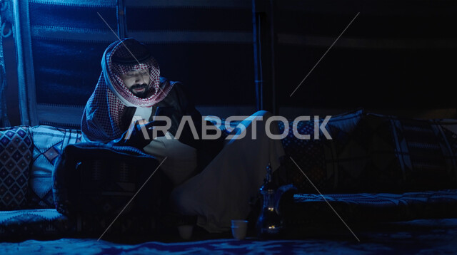 التواصل الاجتماعي عبر تطبيقات الجوال، صوة مقربة لرجل عربي خليجي سعودي يرتدي ملابس الشتاء الدافئة ويحمل بيده الهاتف المحمول مع اضاءة خافته ، التسوق الإلكتروني، جلسة بر عربية شعبية، كشتة بر شبابية مسائية، مخيمات سياحية في الصحراء، اجواء شتوية ليلية