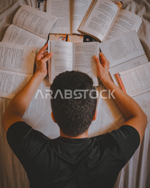 Reading, reading, and preparing for the new academic world. An overhead image from behind of a young Saudi Arabian Gulf man lying down and reading books, scientific books and cultural novels. Love of reading and education, developing mental skills and pra