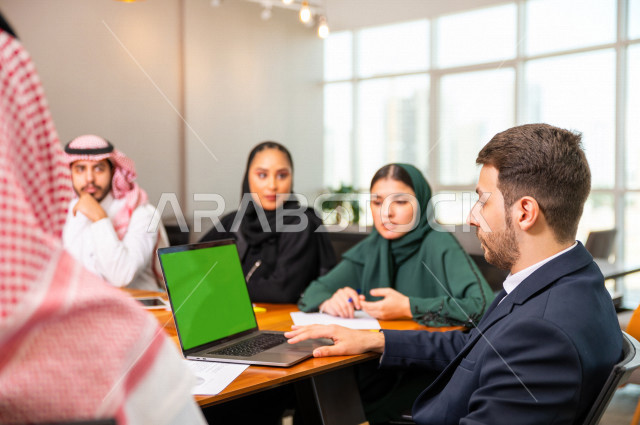 A group of Saudi Gulf businessmen and businesswomen, using laptop technology with a green chroma background, gathered together in the meeting room inside the company's headquarters, a Saudi company, the work environment
