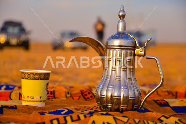 جلسة بر شعبية في صحراء المملكة العربية السعودية، صورة مقربة لدلة قهوة عربية مع كوب ورقي اصفر على سجادة شعبية في المناطق الصحراوية، تقديم المشروبات الساخنة