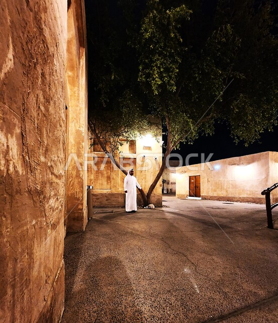 رجل عربي خليجي قطري يرتدي الغترة والعقالويقف في سوق واقف في مدينة الدوحة، الأشجار والنباتات الخضراء ليلا، معالم قطر المشهورة، المباني الطينية التاريخية القديمة، اماكن سياحية في قطر