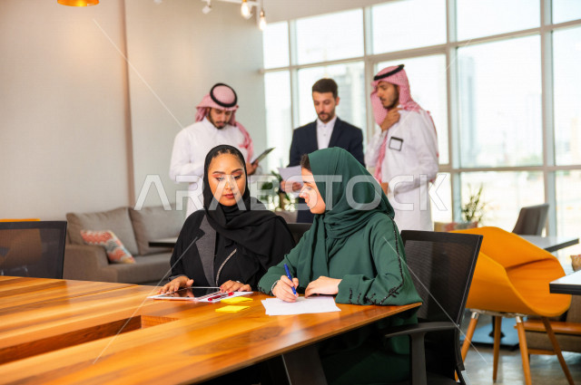 سيدتان اعمال سعوديات خليجيات في غرفة الاجتماعات داخل مقر الشركة ، و بالخلفية مجموعة رياديون من رجال الأعمال السعوديون , شركة سعودية ، شركاء عمل ، بيئة عمل ، وظيفة مكتبية ، عمل خليجي ،  فريق العمل 