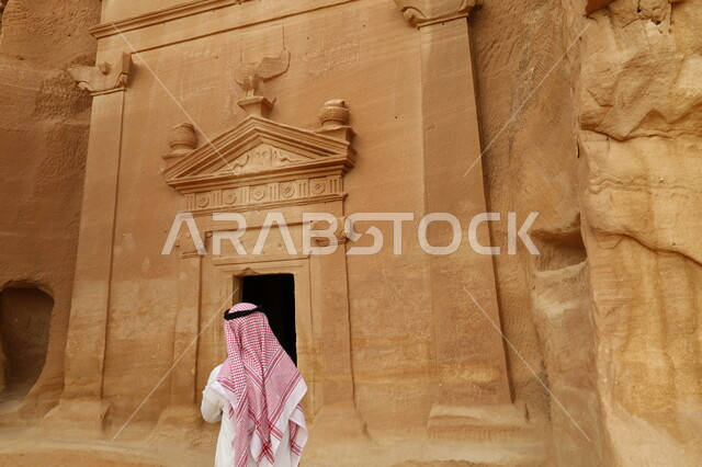 صورة من الخلف لمرشد سياحي عربي خليجي سعودي يقف أمام قصر الفريد في مدائن صالح في محافظة العلا، التكوينات الصخرية الطبيعية في المناطق الصحراوية، المعالم التاريخية الأثرية القديمة في المملكة العربية السعودية، أماكن سياحة في السعودية