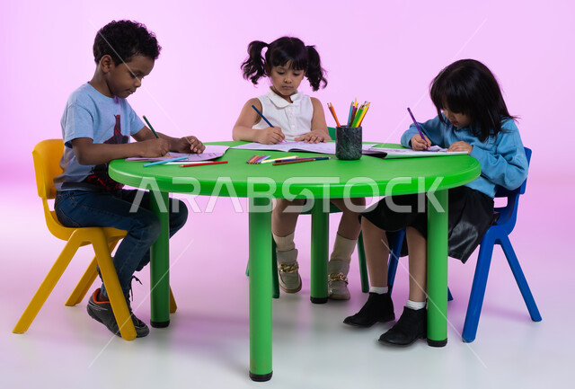 تنمية القدرات العقلية والذهنية بالقيام بالمهارات و الأنشطة المختلفة، الجلسات والحصص التعليمية في الروضات السعودية، الاندماج والتركيز في الرسم والتلوين، بورتريه علوي لأطفال عرب خليجيين سعوديين يرتدون الزي الكاجوال يقومون بنشاط طلاء الرسومات، خلفية وردية.