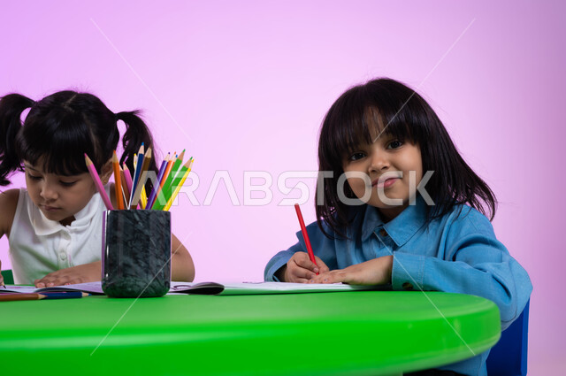 النظر إلى الكاميرا، الحصص الإبداعية في الروضات السعودية، أنشطة تساعد على تنمية القدرات الفكرية، الاندماج والتركيز في الرسم والتلوين، بورتريه مقرب لطفلتين عربيتين خليجيتين سعوديتين ترتديان الزي الكاجوال تبدعان بنشاط طلاء الرسومات، خلفية وردية.