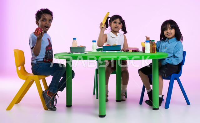 تعليم الأولاد في الروضة أسس الغذاء الصحي السليم لتقوية أجسامهم ونموها، مدارس وحضانات ومراكز ضيافة للأطفال، بورتريه لأطفال عرب خليجيين سعوديين يرتدون الزي الكاجوال يتناولون الطعام الصحي وقت الفسحة المدرسية في إحدى رياض الأطفال في السعودية، خلفية وردية