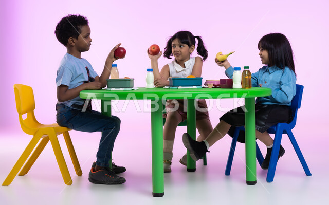 مدارس وحضانات ومراكز ضيافة للأطفال، تعليم الأولاد في الروضة أسس الغذاء الصحي السليم لتقوية أجسامهم ونموها، بورتريه لأطفال عرب خليجيين سعوديين يرتدون الزي الكاجوال يتناولون الطعام الصحي وقت الفسحة المدرسية في إحدى رياض الأطفال في السعودية، خلفية وردية