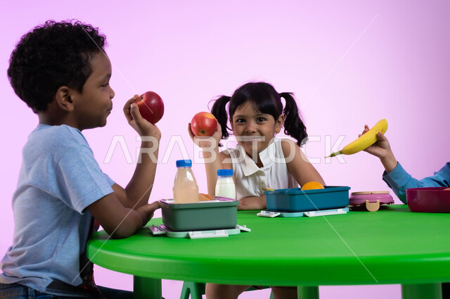 أنشطة لتعليم الأولاد في الروضة أسس الغذاء الصحي السليم، أماكن لحضانة الأطفال والاعتناء بهم وتسليتهم، بورتريه لأطفال عرب خليجيين سعوديين يرتدون الزي الكاجوال يتناولون الطعام الصحي وقت الفسحة المدرسية في إحدى رياض الأطفال في السعودية، خلفية وردية.