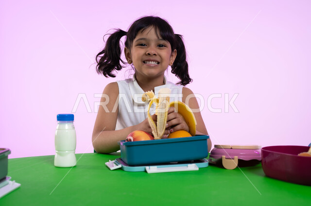 Teaching kindergarten children the basics of proper healthy food, facial gestures indicating happiness, a close-up portrait of a Saudi Arabian Gulf girl in casual clothes looking at the camera eating healthy foods at school break time in a kindergarten in