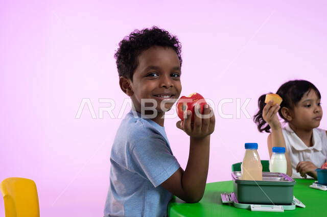 أنشطة لتعليم الأولاد في الروضة أسس الغذاء الصحي السليم، أماكن لحضانة الأطفال والاعتناء بهم، بورتريه لطفل عربي خليجي سعودي يرتدي الزي الكاجوال ينظر إلى الكاميرا يتناول الأطعمة الصحية في وقت الفسحة المدرسية في إحدى رياض الأطفال في السعودية، خلفية وردية