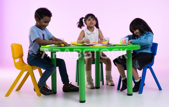 أنشطة فنية تساعد على تعزيز الإبداع والمهارات الحركية والحسية الدقيقة عند الأولاد، الاندماج والتركيز في صنع أشكال باستخدام العجينة الملونة، بورتريه لأطفال عرب خليجيين سعوديين يبدعون بنشاط اللعب بالمعجون الملون في الروضة، خلفية وردية.