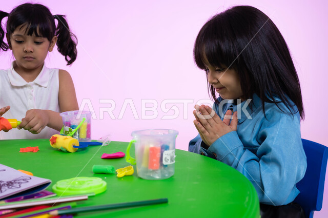 ايماءات الحيرة في اختيار ألوان المعجون، متعة اللعب بعجينة التشكيل الملونة، نشاطات فنية تساعد على تنمية الجانب الإبداعي لدى الأطفال، بورتريه لطفلتين عربيتين خليجيتين سعوديتين ترتديان الزي الكاجوال تقومات باللعب بالصلصال في الروضة، خلفية وردية.