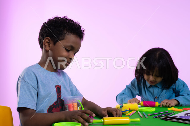 أنشطة فنية وتعليمية تساعد على تعزيز الإبداع والمهارات الحركية والحسية الدقيقة عند الأولاد، الاندماج والتركيز في صنع أشكال باستخدام الصلصال، بورتريه مقرب لأطفال عرب خليجيين سعوديين يبدعون بنشاط اللعب بالمعجون الملون في الروضة، خلفية وردية.