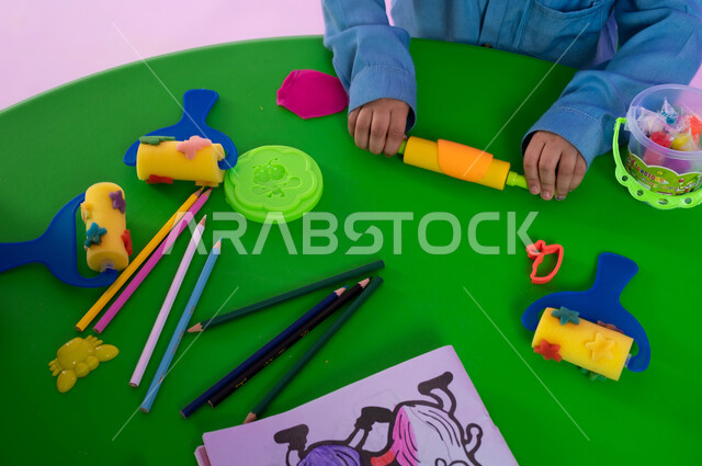 قرطاسية وأدوات مدرسية، متعة اللعب بعجينة التشكيل الملونة، نشاطات فنية تساعد على تنمية الجانب الإبداعي والابتكاري لدى الأطفال، بورتريه علوي مقرب لطفلة عربية خليجية سعودية تقوم باللعب بالصلصال، خلفية وردية.
