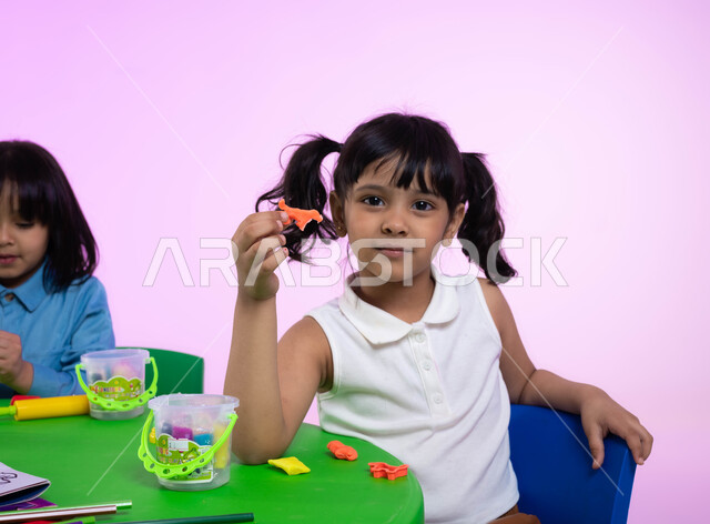 نشاطات فنية تساعد على تنمية الجانب الإبداعي والابتكاري لدى الأطفال، طفلة سعودية تنظر إلى الكاميرا تحمل عجينة ملونة على شكل حيوان، بورتريه لطفلتين عربيتين خليجيتين سعوديتين تبدعان بنشاط اللعب بالصلصال، خلفية وردية.