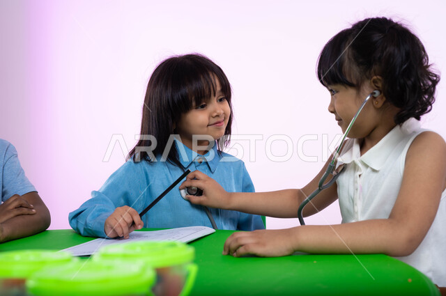 اختيار وتحديد المهنة المستقبلية منذ الصغر ، تعريف الأطفال في الروضة مفهوم الطب والرعاية الصحية، بورتريه مقرب لطفلتين عربيتين خليجيتين سعوديتين تقومان باللعب بممارسة مهنة الطب، طفلة سعودية تقوم بالفحص بواسطة السماعة الطبية، خلفية زهرية.