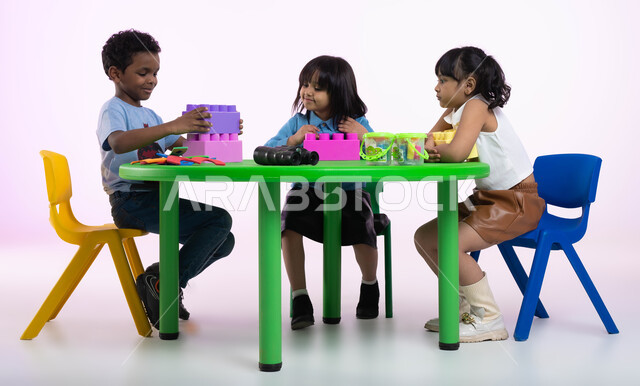 الجلسات والحصص الإبداعية في الروضات السعودية، مهارات وأنشطة تساعد على التطور الفكري، إيماءات وجه تدل على السعادة والمتعة والتركيز، بورتريه لأطفال عرب سعوديين خليجيين يرتدون الزي الكاجوال يستمتعون بتركيب المكعبات، خلفية وردية.