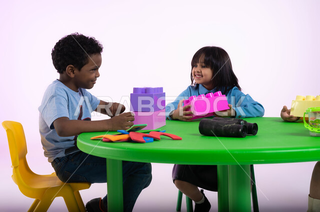 الإبداع وتنمية المهارات والقدرات الذهنية، أماكن لحضانة الأولاد والاعتناء بهم وتسليتهم، إيماءات وجه تدل على السعادة والمتعة، بورتريه مقرب لأطفال عرب سعوديين خليجيين يرتدون الزي الكاجوال يستمتعون بتركيب المكعبات، خلفية وردية.