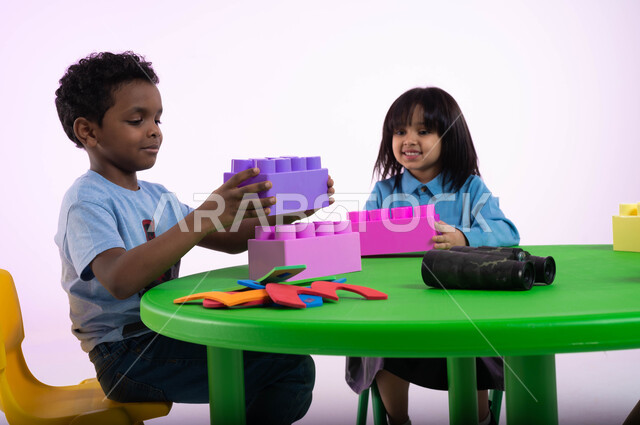 أماكن لحضانة الأولاد والاعتناء بهم وتسليتهم، الجلسات والحصص الإبداعية في الروضات السعودية، إيماءات وجه تدل على السعادة والمتعة والتركيز، بورتريه مقرب لأطفال عرب سعوديين خليجيين يرتدون الزي الكاجوال يستمتعون بتركيب المكعبات، خلفية وردية.