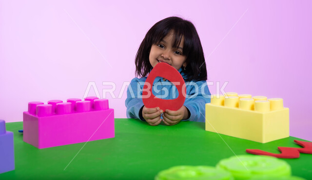 نشاطات ترفيهية وتعليمية للأطفال في الروضة، تعلم الأرقام والأعداد العربية وأشكالها، بوتريه مقرب لطفلة عربية خليجية سعودية ترتدري الزي الكاجوال تنظر إلى الكاميرا بإيماءات تدل على السعادة والثقة بالنفس، خلفية وردية.