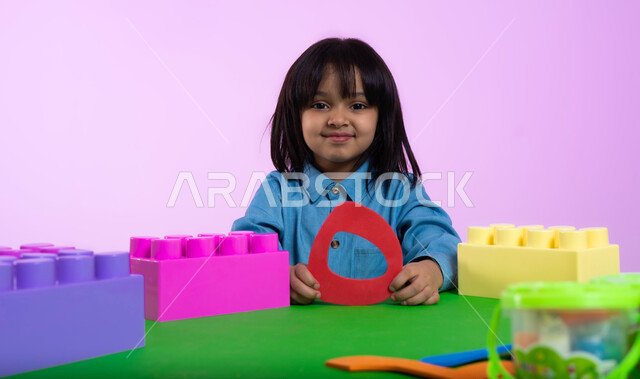 تعلم الأرقام والأعداد العربية وأشكالها، نشاطات ترفيهية وتعليمية للأطفال في الروضة، بوتريه مقرب لطفلة عربية خليجية سعودية ترتدري الزي الكاجوال تنظر إلى الكاميرا بإيماءات تدل على السعادة والثقة بالنفس، خلفية وردية.