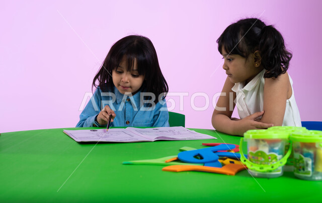 محاولة تعلم النطق الصحيح لمخارج الحروف، طفلة عربية خليجية سعودية تحاول القراءة والتهجئة من كتاب اللغة العربية في الروضة، بورتريه لطفلتين عربيتين سعوديتين خليجيتين ترتديان الزي الكاجوال تتعاونان في حل التمارين، خلفية وردية.