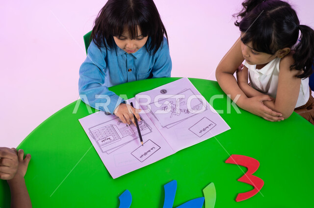 أنشطة تعليمية ولغوية تساعد على تقوية التواصل عند الأولاد، طفلة عربية خليجية سعودية تحاول القراءة والتهجئة من كتاب اللغة العربية في الروضة، بورتريه علوي لأطفال عرب سعوديين خليجيين يرتدون الزي الكاجوال يتعاونون في حل التمارين، خلفية وردية.