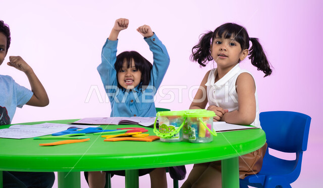 المنافسة والتحدي والتشجيع في النشاطات بين الأطفال في الروضة، بورتريه مقرب لأطفال عرب خليجيين سعوديين يرتدون الزي الكاجوال يرفعون أيديهم، إيماءات يدين تدل على الحماس والشغف، الفرح والسعادة بالفوز والانتصار، خلفية وردية.