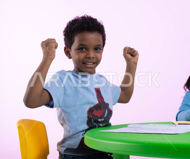 المنافسة والتحدي والتشجيع بين الأطفال في الروضة، بورتريه مقرب لطفل عربي خليجي سعودي يرتدي الزي الكاجوال يرفع يديه مضموم الكفين، إيماءات يدين تدل على الحماس والشغف، الفرح والسعادة بالفوز والإنتصار، النجاح والقوة وتحقيق الأهداف، خلفية وردية.
