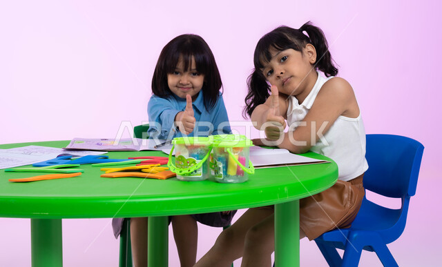 النظر إلى الكاميرا، إيماءات وجه ويدين تدل على الرضا والقبول والإعجاب، أماكن ترفيهية تعليمية للأطفال في المملكة العربية السعودية، بورتريه لطفلتين عربيتين خليجيتين سعوديتين ترتديان الزي الكاجوال تشيران بإصبعهما الإبهامي علامة لايك، خلفية ملونة.