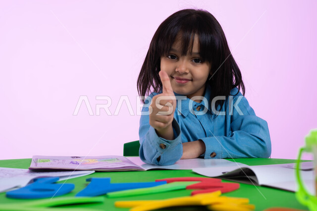 أماكن ترفيهية تعليمية للأطفال في المملكة العربية السعودية، بورتريه لطفلة عربية خليجية سعودية ترتدي الزي الكاجوال تشير بإصبعها الإبهامي علامة لايك، إيماءات تدل على الرضا والقبول والإعجاب، النظر إلى الكاميرا، خلفية ملونة. 