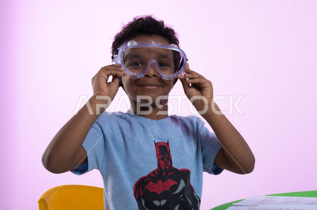 أنشطة ترفيهية وتعليمية للأطفال، بورتريه مقرب لطفل عربي خليجي سعودي يرتدي الزي الكاجوال ونظارات السباحة، الحماس والشوق للغوص في الماء، الاستعداد والتحضير للسباحه، اللعب واللهو بأدوات المسبح، خلفية وردية.
