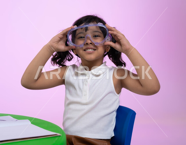 الاستعداد والتحضير للسباحة، بورتريه مقرب لطفلة عربية خليجية سعودية ترتدي الزي الكاجوال ونظارات السباحة، ايماءات الحماس والشوق للغوص في الماء، اللعب واللهو بأدوات المسبح، خلفية وردية.