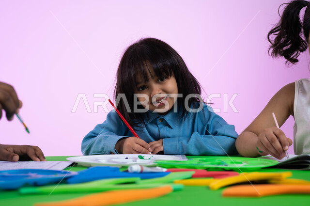 أنشطة ترفيهية وفنية في الروضة، تنمية مهارات وتعليم وإبداع، بورتريه مقرب لأطفال عرب خليجيين سعوديين يرتدون الزي الكاجوال يبدعون بنشاط تلوين الرسومات، إيماءات وجه تدل على السعادة، خلفية وردية.