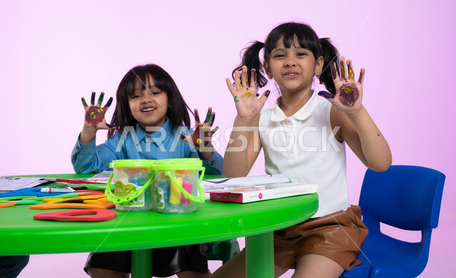 الحصص الإبداعية في الروضات، تقنيات الرسم لمرحلة ماقبل المدرسة، أنشطة تساعد على التطور الفكري، إيماءات تدل على السعادة، بورتريه لطفلتين عربيتين سعوديتين خليجيتين ترتديان الزي الكاجوال تشيران بيديهما، طباعة الألوان المائية بكف اليد، خلفية وردية.