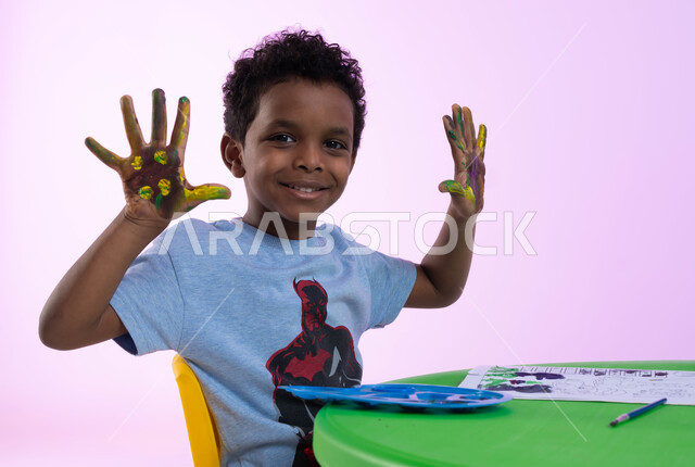 الحصص الإبداعية في الروضات السعودية، تقنيات الرسم لمرحلة ماقبل المدرسة، أنشطة تساعد على التطور الفكري، إيماءات تدل على المتعة والسعادة، بورتريه لطفل عربي سعودي خليجي يرتدي الزي الكاجوال يشير بيديه، طباعة الألوان المائية بكف اليد، خلفية وردية.