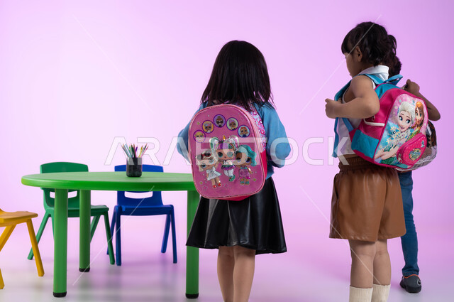 الاستعداد والتجهيز للعودة إلى المنزل، قرطاسية وحقائب مدرسية للطلاب، تنمية مهارات وتعليم وإبداع في الروضات العربية الخليجية السعودية، بورتريه من الخلف لأطفال عرب خليجيين سعوديين يرتدون الزي الكاجوال وحقيبة الظهر، خلفية وردية.