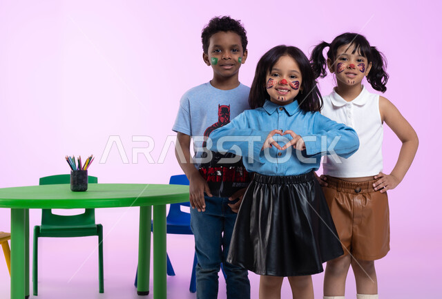 الرسم على وجوه الأطفال، قضاء أوقات ممتعة ومفيدة في الروضة، الشعور بالحب والإعجاب، بورتريه لأطفال عرب خليجيين سعوديين يرتدون الزي الكاجوال ينظرون إلى الكاميرا، إيماءات يدين على شكل قلب، خلفية وردية.