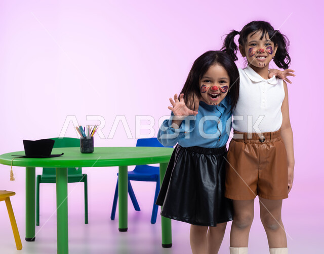 التظاهر والتمثيل بالحيوانات المفترسة، قضاء أوقات ممتعة بالقيام بأنشطة ترفيهية في الروضة، بورتريه لطفلتين عربيتين خليجيتين سعوديتين ترتديان الزي الكاجوال تلعبان وتمرحان، الرسم بألوان صحية على وجوه الأطفال، ايماءات تدل على الفرح والسعادة، خلفية وردية.